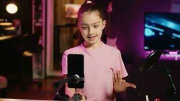 Kind Filme mit Smartphone befestigt zu Selfie Stock im Rosa Neon- zündete Leben Zimmer benutzt wie Fachmann Studio. jung Medien Star erfasst Aufnahmen mit Handy Kamera, diskutieren Über Spaß Tag beim Schule foto