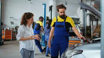 geschickt Mechaniker Portion Klient mit Auto Instandhaltung im Auto Reparatur Geschäft. Mitarbeiter im Garage Einrichtung suchen Über Automobil Teile mit Frau, Ausbessern ihr Fahrzeug während jährlich Inspektion foto