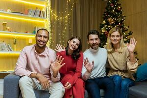 festlich online treffen. ein Gruppe von jung interracial Menschen sind Sitzung auf ein Sofa im ein Wohnung in der Nähe von ein Weihnachten Baum, suchen und winken beim das Kamera, lächelnd, wünsche glücklich Neu Jahr Feiertage. foto