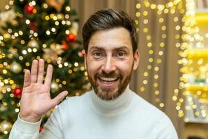 online Schöne Grüße Video Forderung, Weihnachten Mann sieht aus in Netz Kamera und lächelt und Wellen während Sitzung auf Sofa beim Zuhause feiern Neu Jahr Feiertage. foto