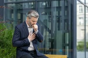 krank reifen Mann draußen Büro Gebäude, Erwachsene grau haarig Geschäftsmann Husten Sitzung auf Bank in der Nähe von arbeiten, Senior Boss im Geschäft passen mit kalt Virus. foto