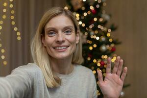 online Video Grüße, Frau beim Zuhause allein auf Weihnachten Ferien feiern Neu Jahr, suchen beim Smartphone Kamera und winken Hand, Video Anruf zu Freunde, Aufzeichnung Sitzung Zuhause in der Nähe von Weihnachten Baum. foto