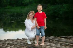 glücklich Mutter und Sohn Stand durch das Fluss auf ein hölzern Brücke, Pose und aussehen beim das Kamera. foto