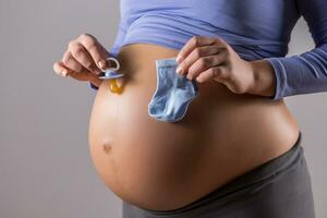 Bild von Bauch von schwanger Frau mit halten Schnuller und Socken zum Baby Junge auf grau Hintergrund. foto