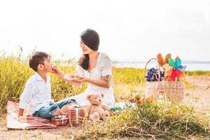 schöne asiatische mutter, die ihrem sohn auf der wiese einen snack füttert, wenn sie picknick macht. Mutter und Sohn spielen zusammen. am Muttertag feiern und das Konzept schätzen. Sommer-Menschen und Lifestyle-Thema foto