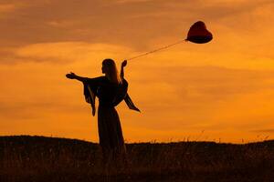 Silhouette von ein Frau halten Herz geformt Ballon und suchen beim schön Sonnenuntergang. foto