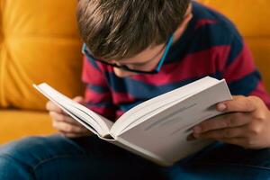 neugierig Schulalter Junge mit Brille lesen ein Buch auf das Couch foto