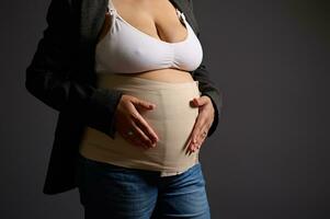abgeschnitten Aussicht von ein jung Frau tragen elastisch Binde auf ihr postnatal Bauch, lächelnd suchen beim Kamera, grau Hintergrund foto