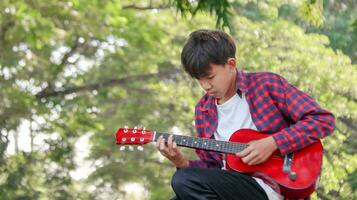 jung asiatisch Junge ist spielen Gitarre im ein lokal Park, Sanft und selektiv Fokus foto