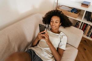 schwarze junge Frau mit Handy beim Ausruhen auf dem Sofa