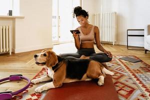 Schwarze junge Frau mit Handy beim Sitzen mit ihrem Hund auf der Matte foto