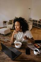 Schwarze junge Frau in Kopfhörern mit Laptop und Frühstück