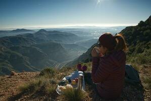Frau nimmt ein brechen auf das oben von das Berg beobachten das Landschaft, mit ein Thermosflasche und ein Tasse von Kaffee im ihr Hand. foto