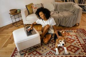 Schwarze Frau, die Laptop benutzt und ihren Hund streichelt, während sie auf dem Boden sitzt foto