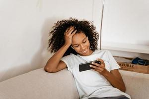 Schwarze junge Frau in Kopfhörern mit Handy beim Ausruhen auf dem Sofa