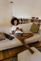 Schwarze junge Frau in Kopfhörern mit Handy beim Ausruhen auf dem Sofa