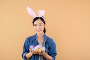 Porträt jung asiatisch Frau tragen Denim Kleider und Hase Hase Ohren und halten bunt Eier isoliert auf Beige Hintergrund. Lebensstil glücklich Ostern Konzept. foto