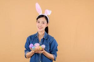 Porträt jung asiatisch Frau tragen Denim Kleider und Hase Hase Ohren und halten bunt Eier isoliert auf Beige Hintergrund. Lebensstil glücklich Ostern Konzept. foto