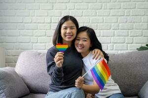 attraktiv asiatisch Lesben Paar halten lgbt Flagge im Leben Zimmer beim Zuhause mit lächelnd Gesichter foto