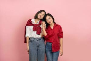 heiter jung asiatisch Lesben Paar im Hemden und Jeans steht halten Hände zu ausdrücken ihr Liebe auf ein Rosa Hintergrund. foto