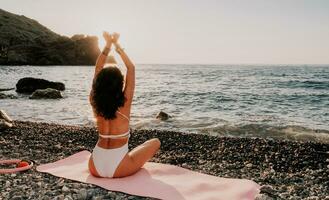 Frau Meer Yoga. glücklich Frau im Weiß Badeanzug und Boho Stil Armbänder üben draußen auf Yoga Matte durch Meer auf Sonnenuntergang. Frauen Yoga Fitness Routine. gesund Lebensstil, Harmonie und Meditation foto