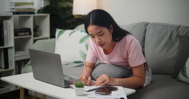 Porträt von jung asiatisch Frau prüft ihr Finanzen mit ein Taschenrechner auf Smartphone und macht Anmerkungen im ein Notizbuch während Sitzung auf das Sofa beim Zu Hause zu Hause Finanzen foto