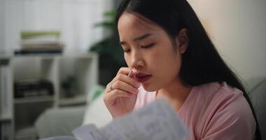 Porträt von jung asiatisch Frau konzentriert im rechnen Budget Sitzung auf Sofa beim Zuhause halten Papier Steuern Rechnungen und lesen finanziell Informationen, Startseite Finanzen foto