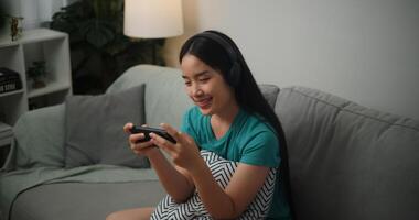 Porträt von jung asiatisch Frau tragen Brille und Kopfhörer genießt spielen online Esport Spiele auf Smartphone Sitzung auf Sofa im das Leben Zimmer beim Zuhause, Gamer Lebensstil Konzept. foto