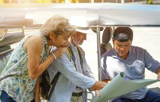 Nahansicht amerikanisch Senior Tourist Mann mit seine Freund europäisch Fragen das Tuk Tuk Thailand Taxi Treiber zum Sie Ziel auf das Karte mit Sonne hell Hintergrund. Senior Tourist Konzept foto