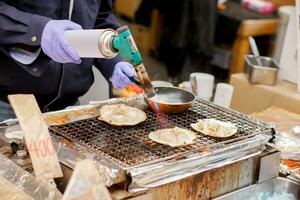 Hand des Küchenchefs beim Brennerkochen und Verbrennen großer Jakobsmuscheln mit Käse auf dem Herd zum Verkauf für Kunden auf dem Kuromon-Markt. scallops bar-bq ist beliebt bei touristen, die nach osaka, japan, kommen. foto