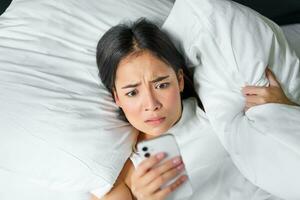 Porträt von asiatisch Mädchen wacht auf oben im Morgen, sieht aus beim Handy, Mobiltelefon Telefon mit schockiert Gesicht, verschlafen, umarmen Kissen und verzog das Gesicht foto