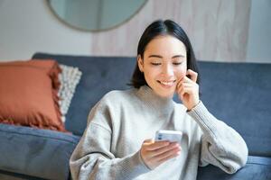 Porträt von lächelnd asiatisch Frau suchen fasziniert beim Smartphone Bildschirm, interessiert mit etw auf Handy, Mobiltelefon Telefon, Sitzung schüchtern auf Fußboden im Leben Zimmer foto