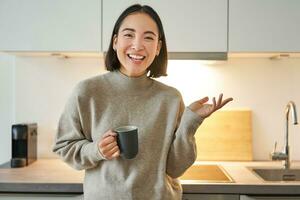 Porträt von stilvoll asiatisch Frau Stehen im Küche mit Becher, Trinken Kaffee, erklären etw, zeigen Banner oder Werbung foto