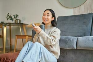 Porträt von jung Koreanisch Frau Aufpassen Fernsehen, halten Fernbedienung und suchen erstaunt beim Fernseher Bildschirm, Ausgaben Zeit beim Zuhause foto