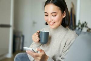 Porträt von lächelnd asiatisch Mädchen Überprüfung ihr Nachrichten Futter auf Smartphone und Trinken Kaffee, Sitzung auf Sofa beim heim, Surfen auf Handy, Mobiltelefon Telefon, lesen foto