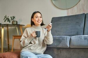 Porträt von lächelnd Koreanisch Frau Sitzung in der Nähe von Fernseher, halten Fernbedienung und Schalten Kanäle während Trinken heiß Kaffee foto