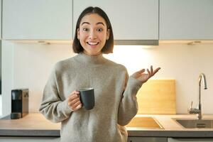 Porträt von stilvoll asiatisch Frau Stehen im Küche mit Becher, Trinken Kaffee, erklären etw, zeigen Banner oder Werbung foto