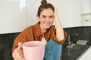 schön jung Brünette Frau sitzt im Küche, Getränke Kaffee, bietet an Sie Tasse und lächelt, sieht aus glücklich foto