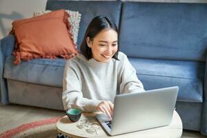 schön asiatisch Mädchen funktioniert von heim, studieren mit Laptop, Sitzung im Leben Zimmer, Sein auf Fernbedienung, freiberuflich tätig online foto