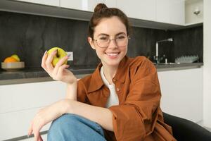 Porträt von lächelnd, modern Frau im Gläser, Sitzung beim Zuhause und Essen Grün Apfel, haben ein Mittagessen brechen, beißen Obst foto