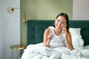 schön jung Koreanisch Frau Berufung, haben ein Telefon Anruf im Bett, reden mit Freund während bleiben im Schlafzimmer foto