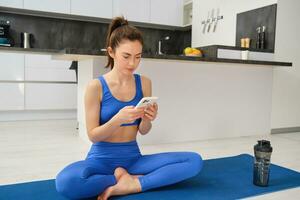 Porträt von Frau mit Fitness Anwendung, tun trainieren von heim, sitzt auf Yoga Matte mit Smartphone im Leben Zimmer foto