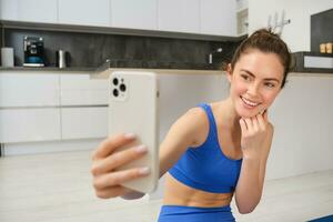 Frau tun trainieren Ausbildung von Zuhause und nehmen Selfie auf Smartphone Kamera, posieren zum Foto Innerhalb ihr Haus, sitzt auf Gummi Yoga Matte im Blau Gamaschen und sportbra