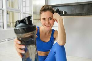 lächelnd Fitness Mädchen Getränke Wasser im das Mitte von Ausbildung Sitzung, lächelnd und suchen Glücklich, trainieren von Zuhause im Blau sportbra foto