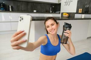 Wohlbefinden und Sport. schön jung Frau nehmen Selfie auf Smartphone, tun Fitness Ausbildung von heim, trinken Wasser, bleibt hydratisiert und tun Übungen foto