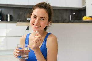 schließen oben Porträt von Brünette Fitness Frau, nehmen Diät Ergänzungen, Vitamine und Glas von Wasser nach trainieren beim Zuhause foto