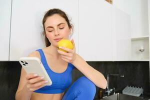 Bild von stilvoll Sportlerin, jung Fitness Lehrer Sitzung im Küche und Essen ein Apfel, halten Smartphone, mit Sozial Medien App auf Handy, Mobiltelefon Telefon foto