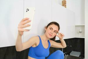 Porträt von lächelnd, schön Fitness Lehrer, posieren zum Fitness Blog, Selfie auf Smartphone, Sitzung im Küche, suchen glücklich foto