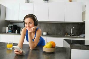 Lebensstil und trainieren. jung lächelnd Frau im Kopfhörer, Stehen im Küche mit Smartphone, Trinken Orange Saft und Hören Musik, Überschrift Über zu Fitnessstudio, gehen Joggen foto