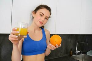 schließen oben Porträt von Sportlerin, Fitness Mädchen halten Glas von frisch Saft und ein Orange im Hände, lächelnd beim Kamera, Stehen im Küche foto